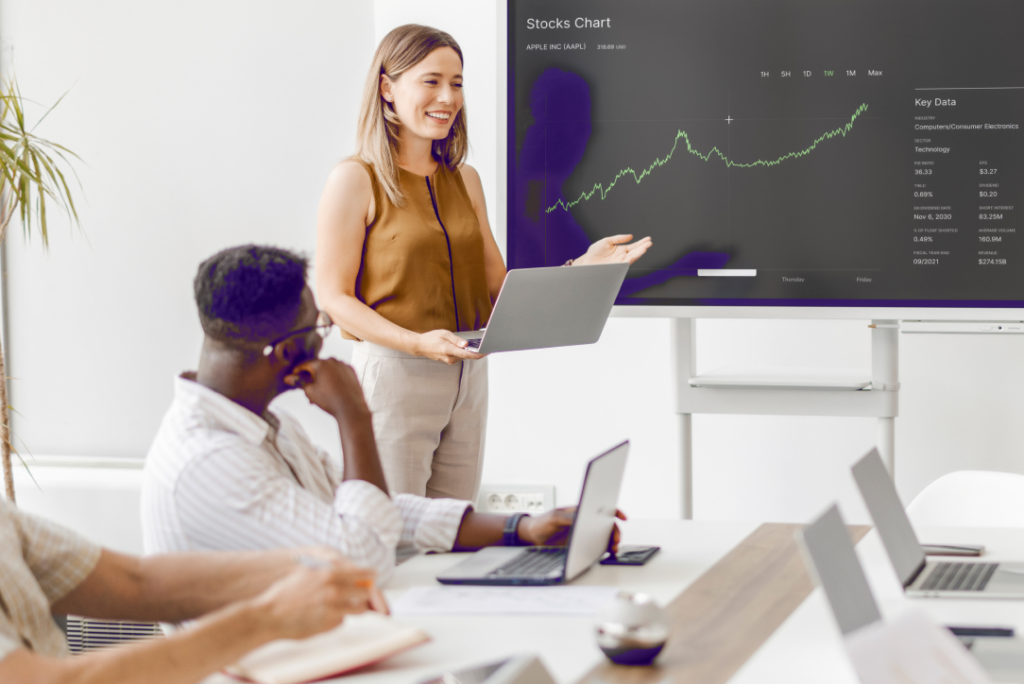 Woman presents stock trends to attentive men in a modern, tech-equipped office setting.