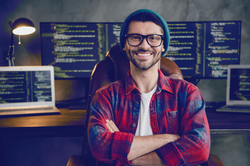 Confident software developer in casual attire smiles in a tech-enhanced office with code on monitors.