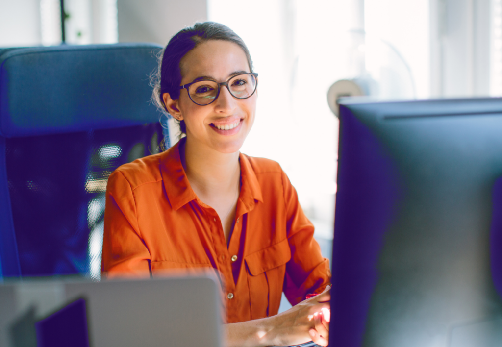 Professional woman in orange blouse working happily at a tech-equipped, modern office desk.